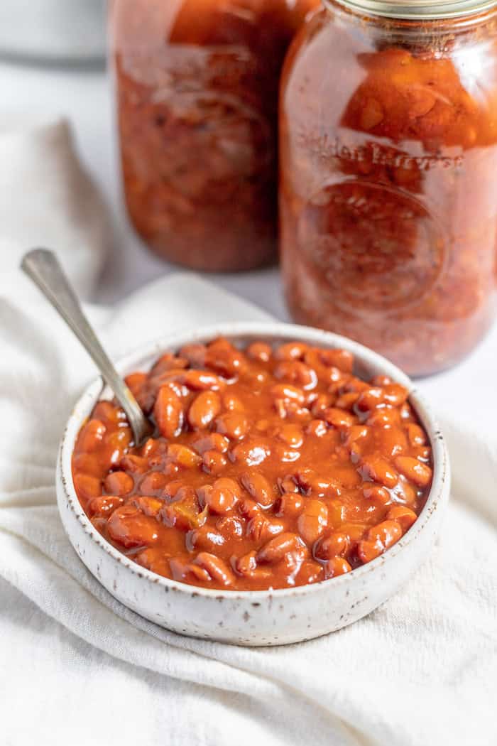 Image shows a bowl of home-canned baked beans with jars of beans in the background on a white cloth