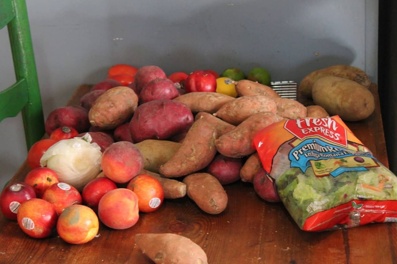 Image shows a table full of fresh produce- apples, peaches, sweet and Russet potatoes, and a bag of salad mix. 