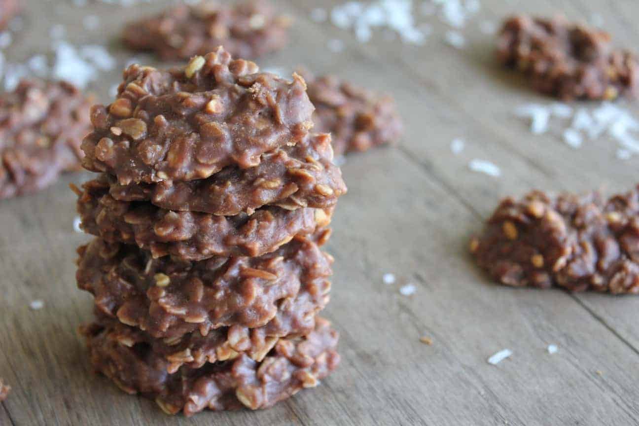 Image shows a stack of chocolate coconut no bake cookies on a countertop. Nearby are several single cookies also placed on the counter, with coconut sprinkled around them. 
