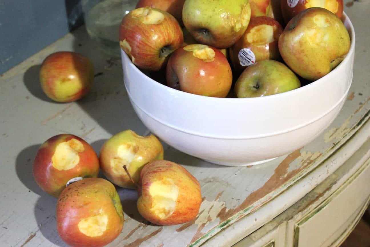 Image shows a large white bowl on a dresser, filled with apples. Each of the apples has a small bite or two taken out of it.