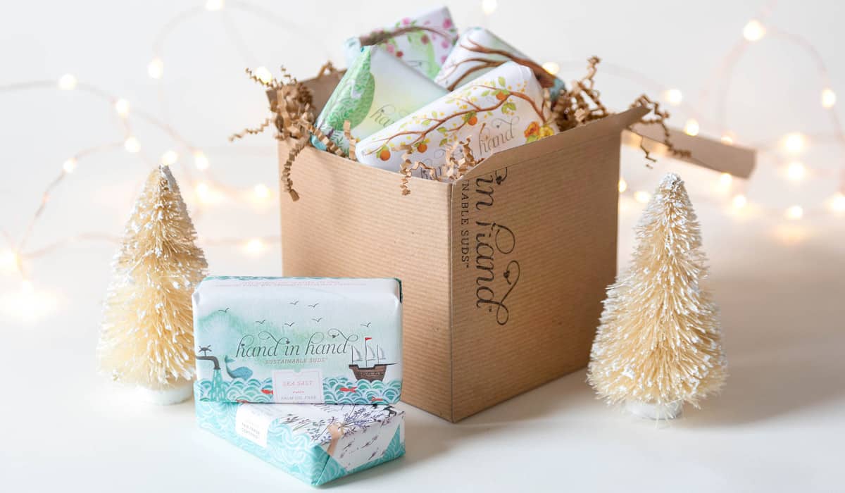 Image shows a gift box from Hand in Hand on a white surface. Handmade soaps sit in front of the box, along with two small tree ornaments