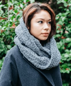 Image shows a young woman with dark hair standing in front of greenery wearing a large cowl neck scarf.