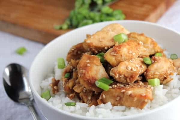 Image shows a bowl of white rice with sesame chicken over it and a spoon resting next to the bowl.