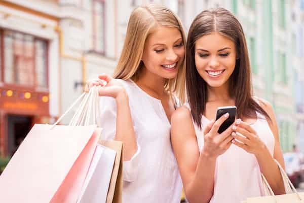 Image shows two woman standing on a street looking at a phone and smiling. One of them is holding several shopping bags.
