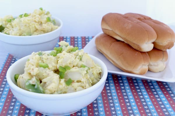 Image shows two bowls of potato salad on a table next to a plate of hot dog buns