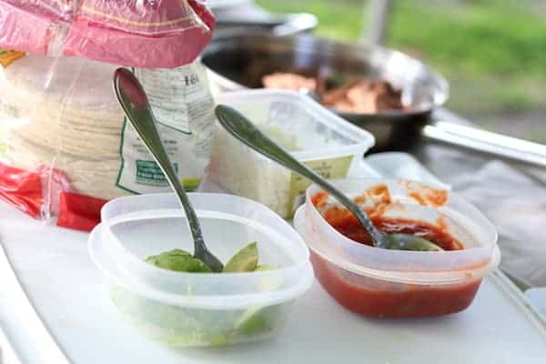 Image shows two Tupperware containers on a table outdoors with salsa and avocados in them. A sleeve of tortilla shells sits nearby.