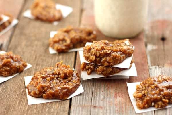 Image shows about a half dozen healthy no-bake pumpkin spice cookies on white parchment paper spread out on a table. A glass of milk sits in the background.