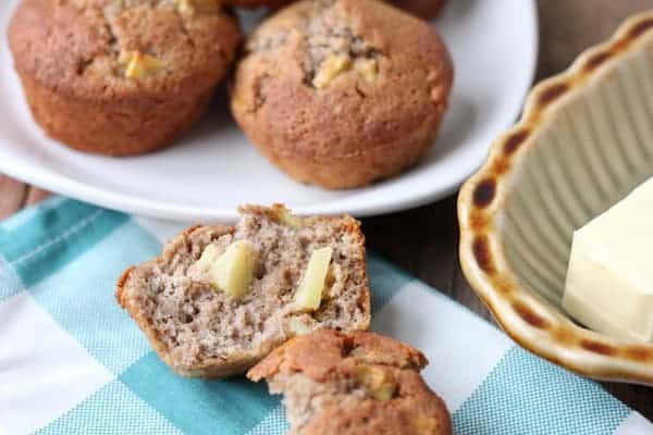 Image shows several apple cinnamon muffins on a plate, with another muffin split open with melting butter on it.