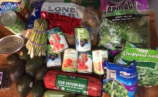 Image shows a table covered in groceries for the week, including cans of vegetables, some fresh vegetables, avocados, asparagus, potatoes, ground beef and more.