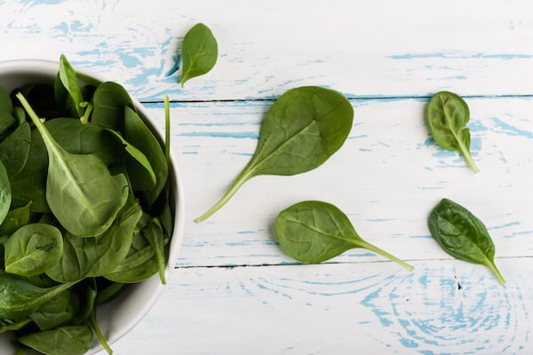 Image, taken from above, shows a bowl of spinach with several spinach leaves scattered on the table next to it