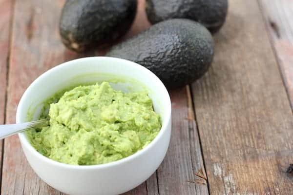 A small bowl of homemade guacamole sits on a table with a spoon in it. Three avocados sit nearby on the table.