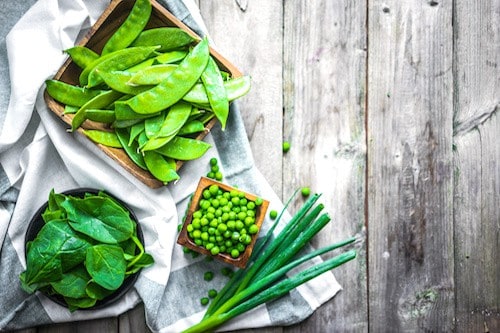 Image, taken from above, shows several green vegetables on a table- snap peas, spinach, green onion.