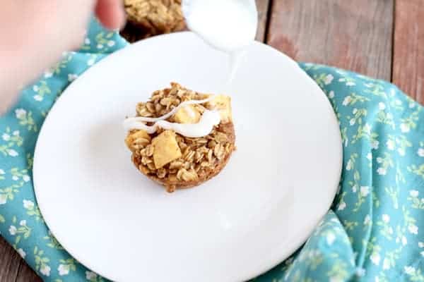 Image shows a hand drizzling some icing over the top of a baked oatmeal cup on a white plate with a printed turquoise towel under it.
