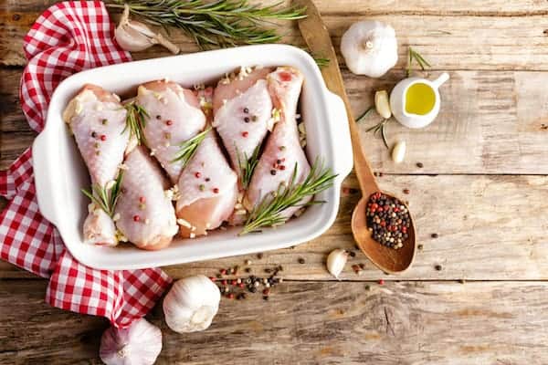 Image, taken from above, shows a white casserole dish with chicken legs in it, rosemary and peppercorns are sprinled on top. Next to the dish sits garlic cloves, and a small container of oil. 