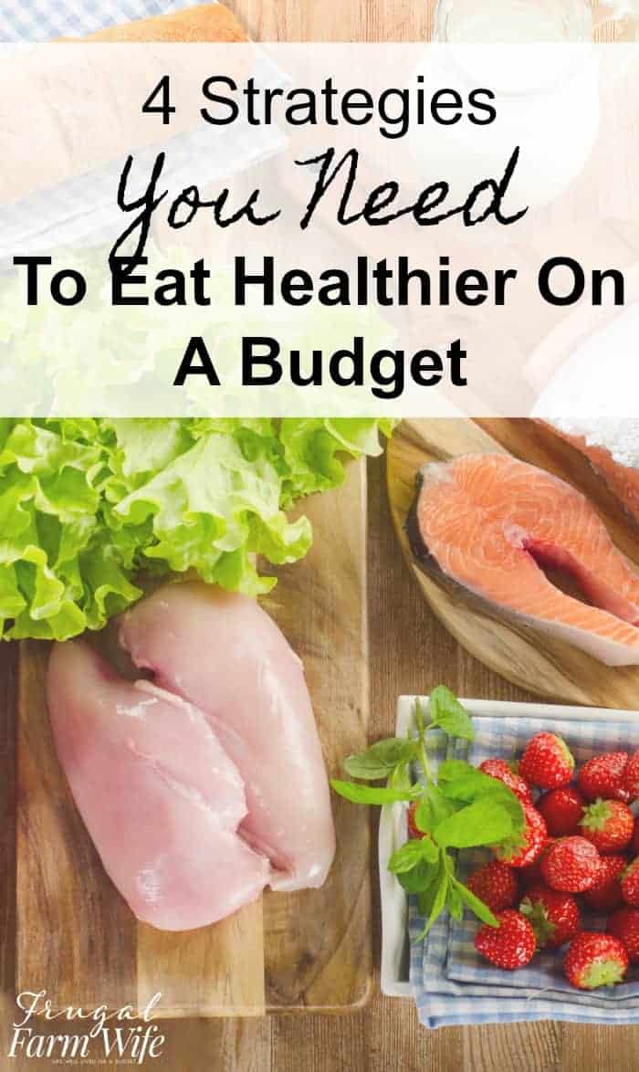 Image, taken from above, shows a counter top with a raw chicken breast and salmon sitting on it, with some lettuce and strawberries nearby. Text overlay reads "4 Strategies You Need to Eat Healthier On a Budget"