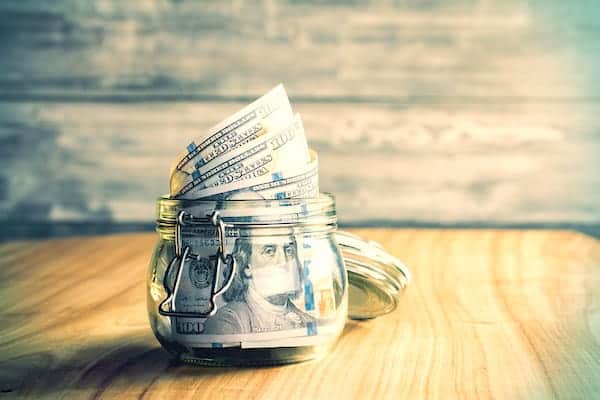 Image shows a small jar full of $100 bills sitting on a wood table.