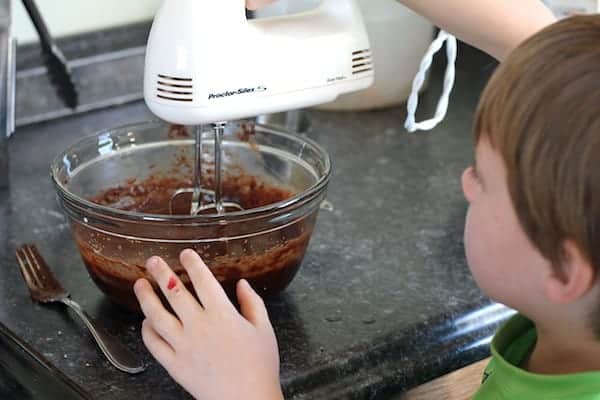 Image shows a small child mixing brownie batter in a bowl with a hand mixer.