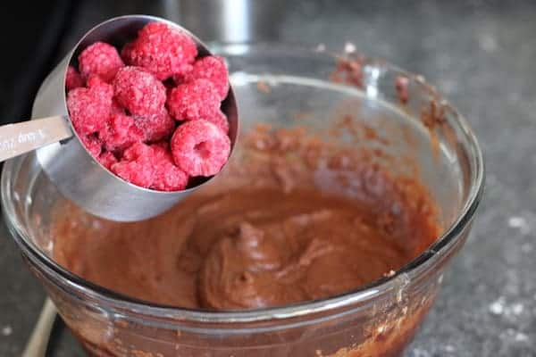 Image shows a bowl of brownie batter with a scoop of raspberries over it about to be poured into the batter.
