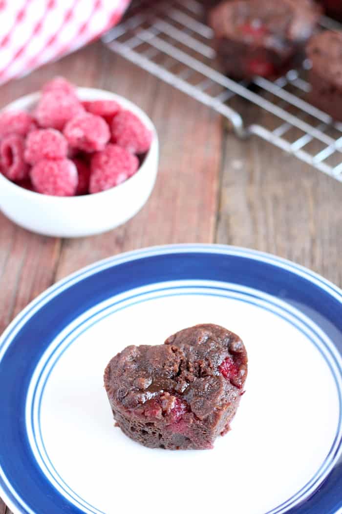 Image shows a chocolate brownie in the shape of a heart on a plate next to a bowl of raspberries