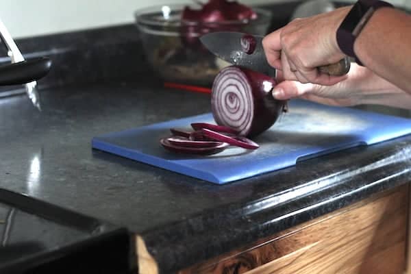 Image shows a pair of hands slicing a red onion on a blue cutting board. 
