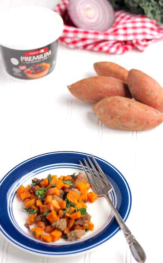 Image shows a small white plate with blue trim of sweet potato hash with a fork. Nearby on the table sit some sweet potatoes and a red and white napkin.
