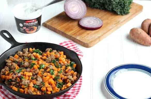 Image shows a large cast iron skilled full of sweet potato hash on a red and white towel. A small white plate with blue trim sits nearby, along with a cutting board, onion, sweet potato and kale. 