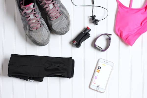 Photo, taken from above, shows must have running items laid out on a white surface- including sturdy shoes, ear buds, mace, a watch, sportsbra, phone and belt bag.