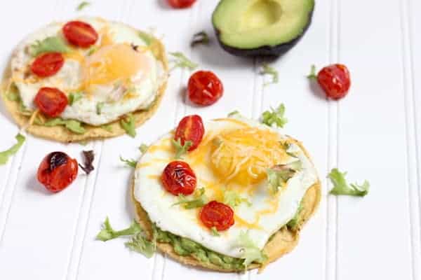 Images shows two simple breakfast tostadas on a counter- topped with egg, avocado, tomato and cheese. several tomatoes and half an avocado sit nearby.