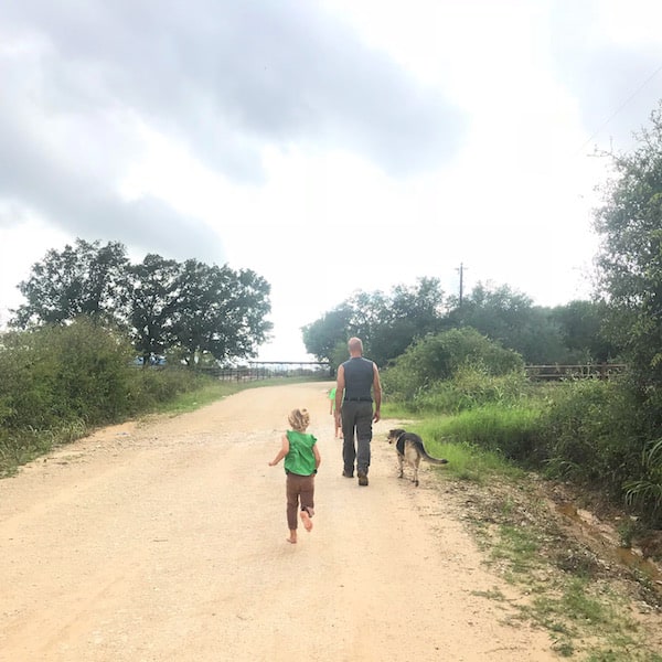 Image shows a man, dog, and small child walking down a dirt road with clouds overhead.