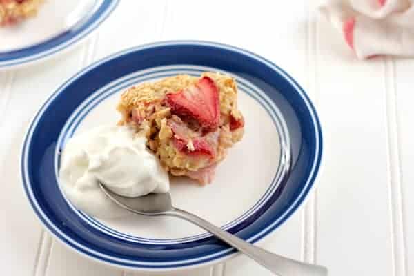 Image shows a white plate with blue trim with a piece of baked strawberry oatmeal on it. A spoon with Greek yogurt sits next to the oatmeal.
