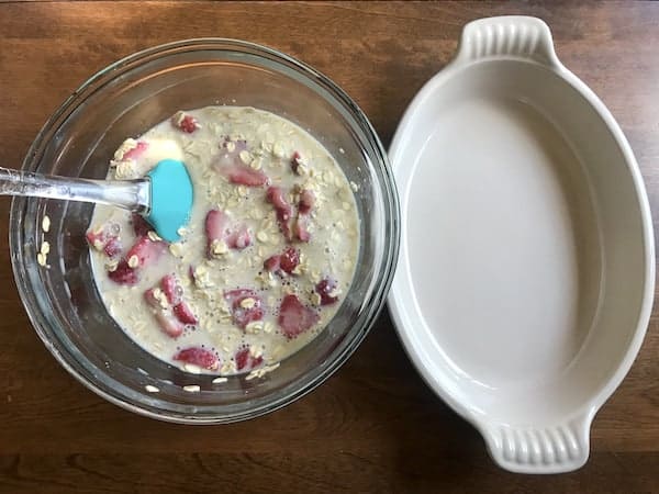 Image, taken from above, shows a white oval pan with a glass bowl of baked oatmeal batter sitting next to it. A spatula sits in the bowl ready to pour the batter into the pan.