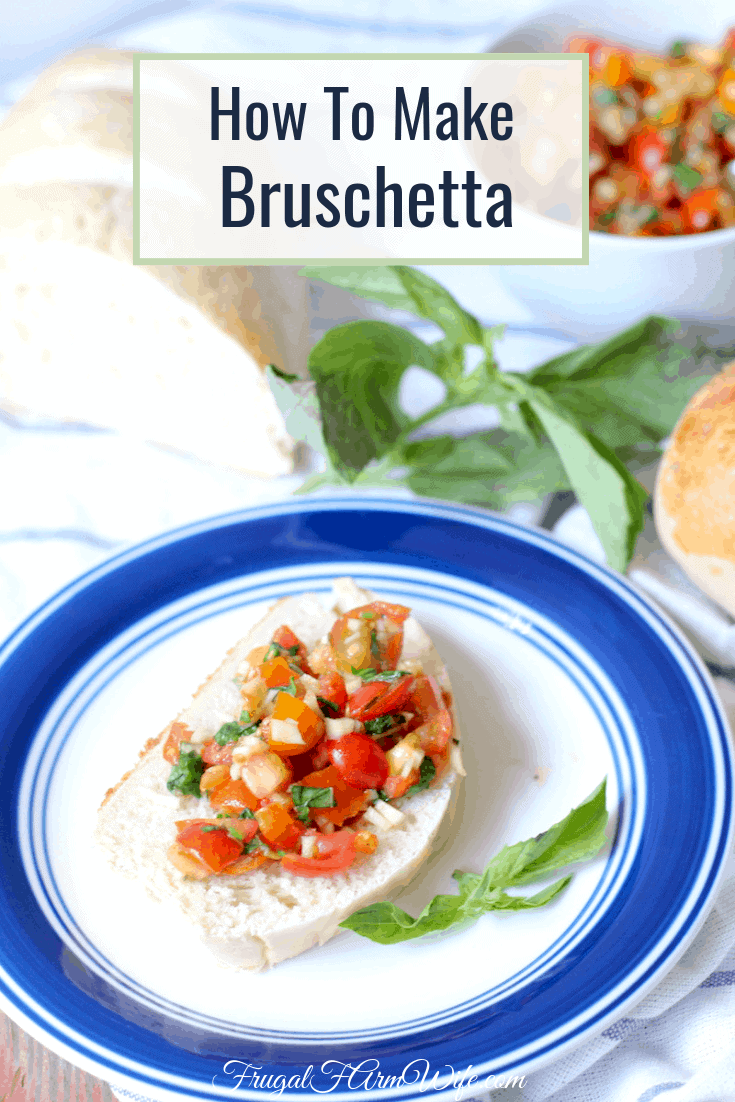 Image shows a small white plate with blue trim on a table with a slice of bread and homemade buschetta on top. A larger bowl of bruschetta sits in the background. 