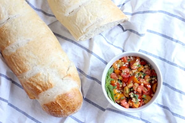 Image shows a bowl of homemade bruschetta on a white and blue striped towel, next to two loaves of crusty bread.