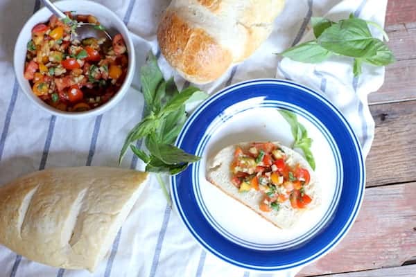 Image, taken from above, shows a white plate with blue trim with a slice of bread topped with homemade bruschetta. A bowl of bruschetta and loaf of homemade bread sit nearby on the table. 