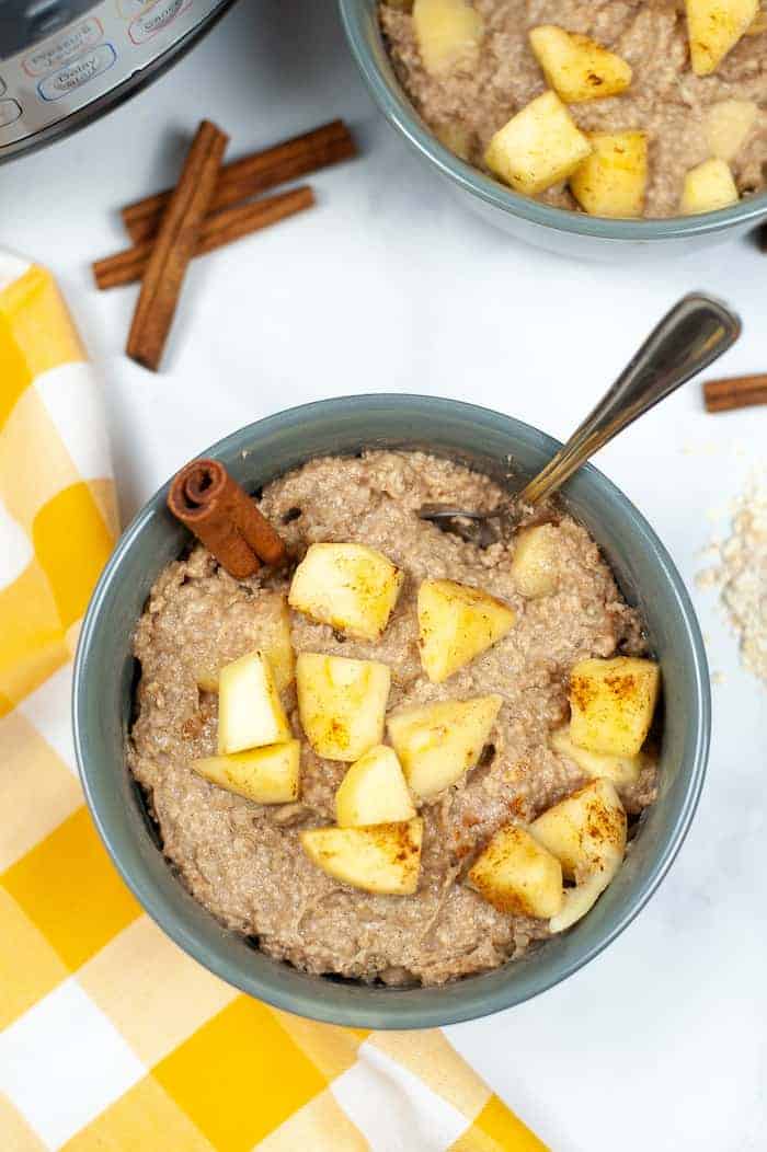 Image shows a bowl of apple cinnamon oatmeal from above, with cinnamon sticks and slices of apples garnishing it