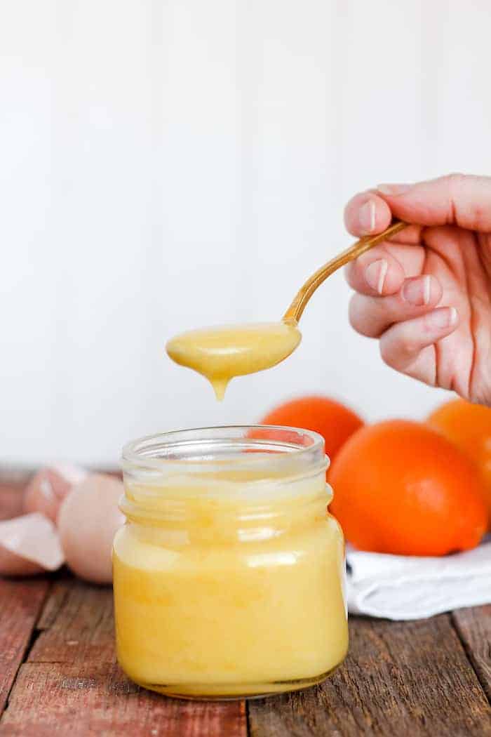 Image shows a hand holding a spoonful of orange curd suspended over a jar