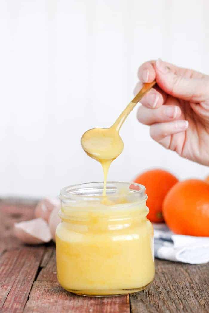 Photo shows a hand holding a spoonful of orange curd over a jar, pouring it back in
