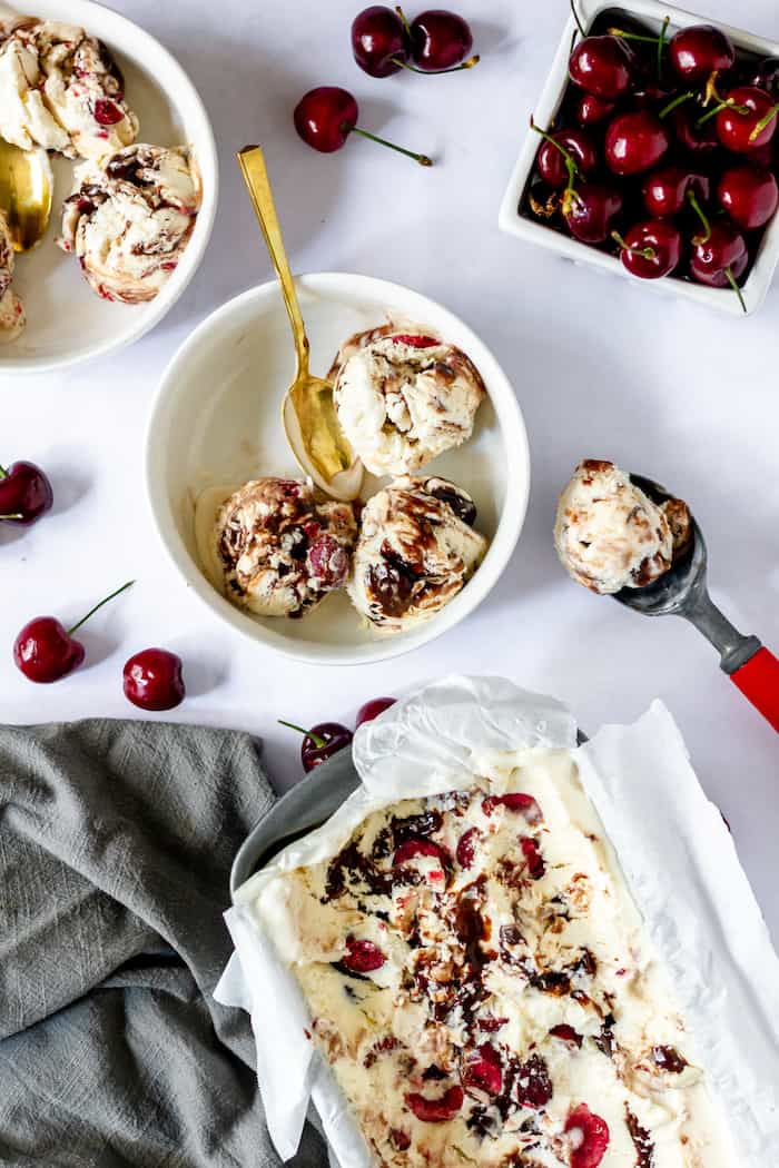 Image, taken from above, shows a bowl of cherry chocolate ice cream on a table with a gold spoon in it. Nearby sits an ice cream scoop with ice cream in it, and a bowl of cherries.