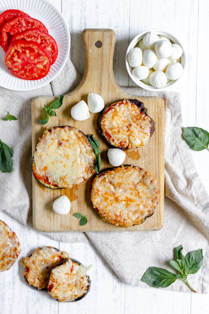 Image shows a cutting board with three small eggplant pizzas, next to a plate of tomato and a bowl of fresh mozzarella