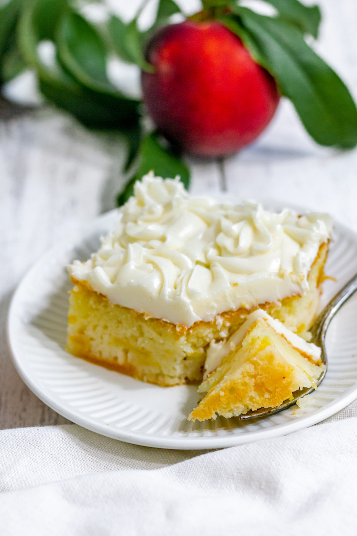 Image shows a close up of a piece of peach cake on a white plate next to a fresh peach.