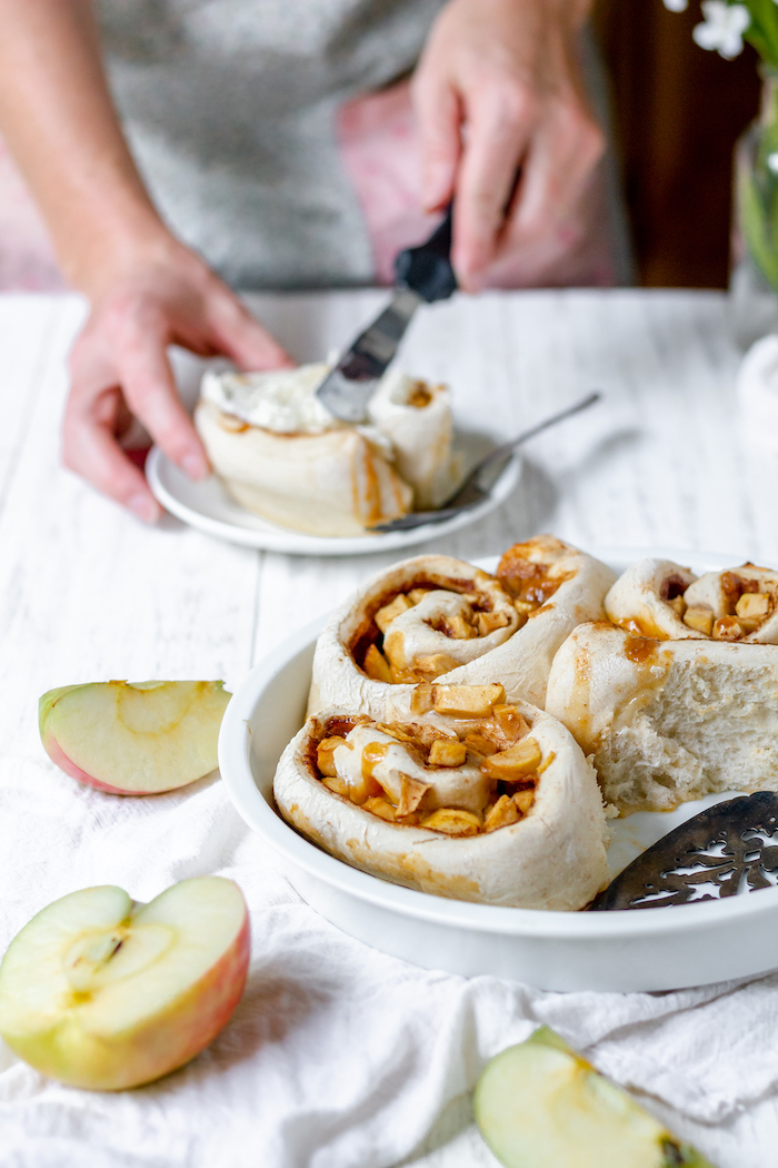 Image shows a hand spreading icing on apple cinnamon rolls