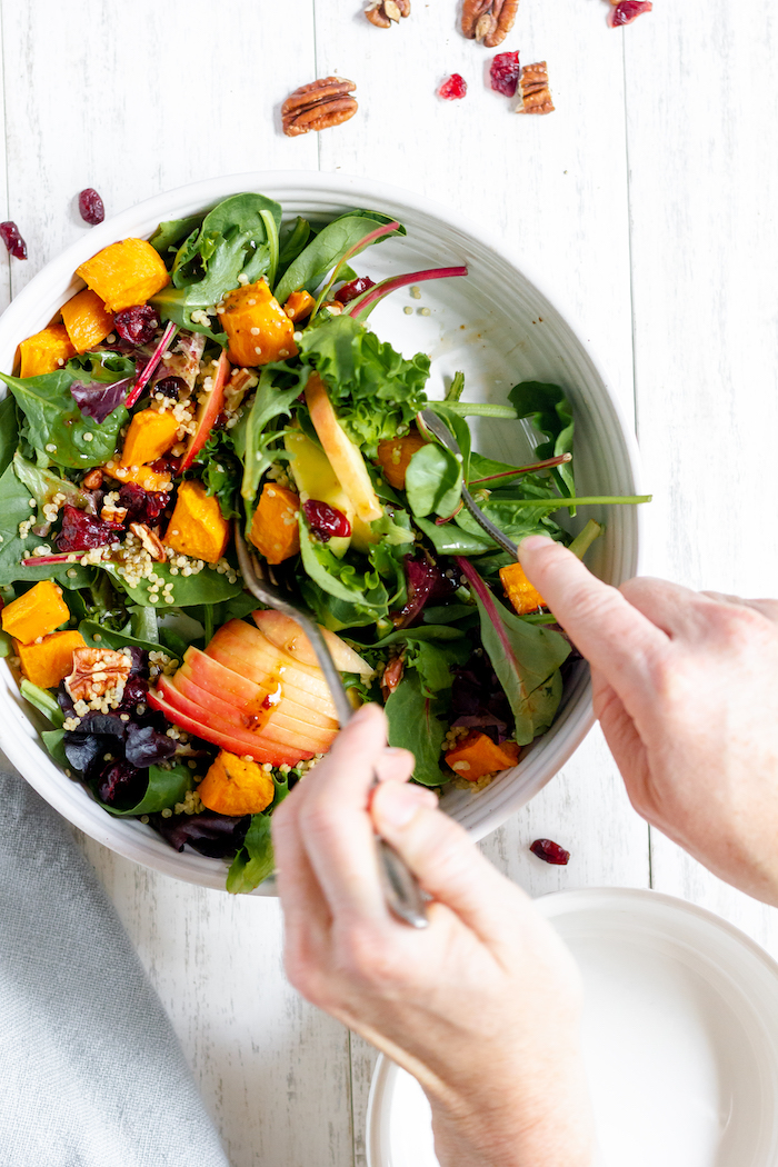 Image shows two hands tossing a fall harvest salad in a bowl