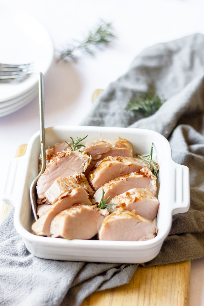 Image shows a casserole dish of Sous Vide Rosemary Pork Loin sliced in it, sitting on a grey tablecloth on a dining table.