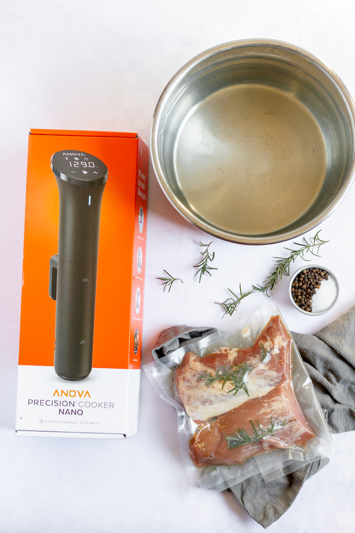 Image, taken from above, shows a large pot, with an Anova Precision Cooker Nano in the box. Next to it sits a wrapped pork loin with salt, pepper, rosemary and a grey hand towel.