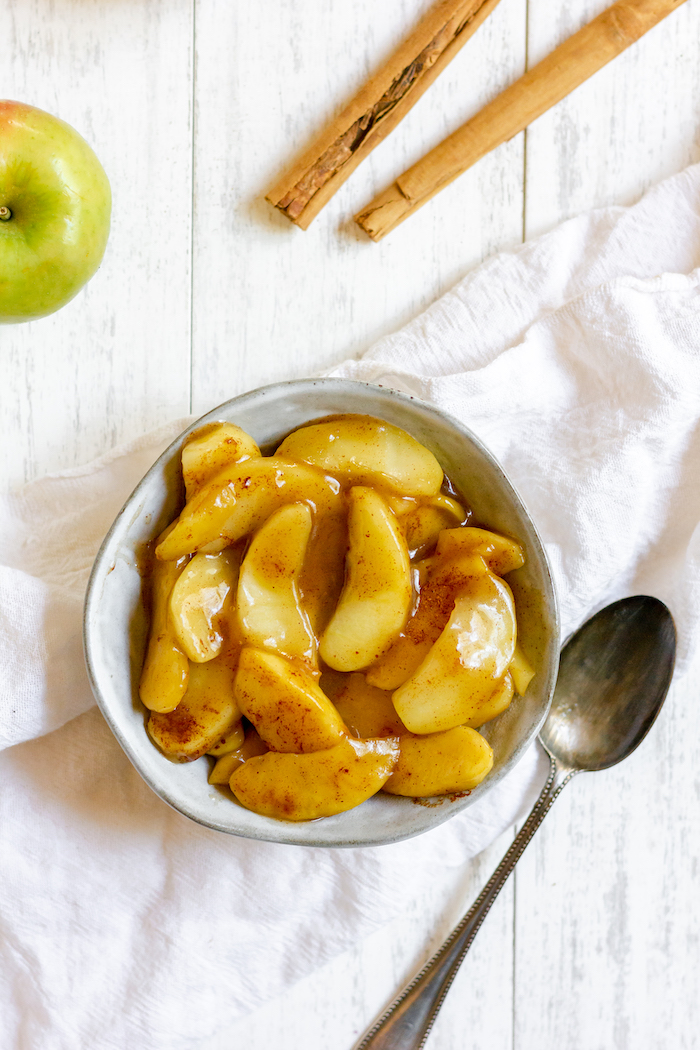 Image shows a bowl of fried apple slices next to some cinnamon sticks and a spoon
