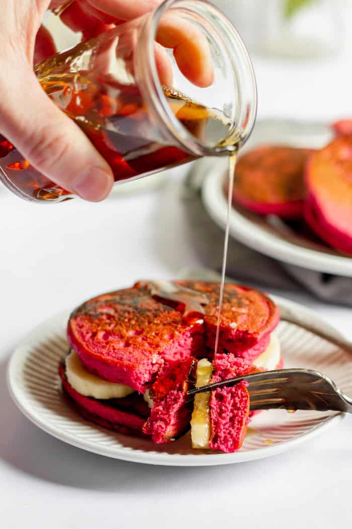 Photo shows a hand pouring syrup over pink beet pancakes