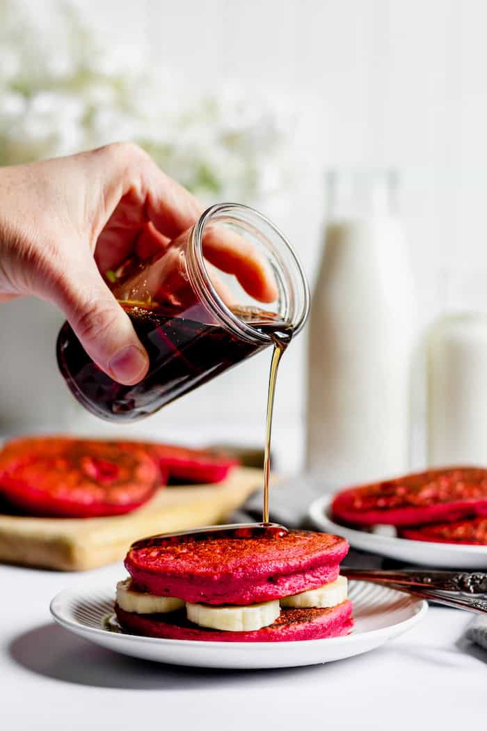 Image shows a hand pouring syrup over some beet pancakes on a plate