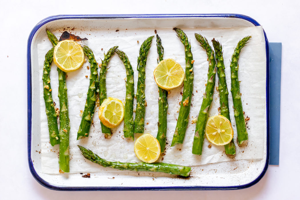 Image, taken from above shows the asparagus stalks on a tray with lemon and garlic, ready to be roasted.