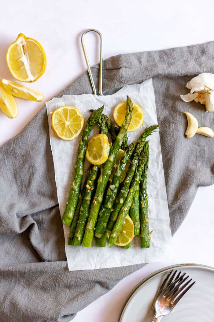 Image, taken from above shows the garlic and lemon asparagus vegetable side dish on a grey towel. Several stalks of asparagus are coated in garlic and lemons.