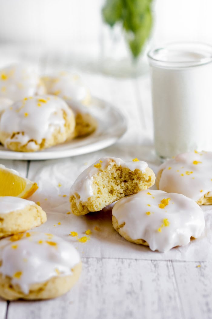 Image shows three iced Italian lemon cookies on a white table. Behind them sits a tall glass of milk, and a plate of more cookies.